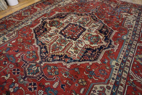 Densely patterned red rug with complex geometric motifs in navy, cream, and teal on wood flooring.