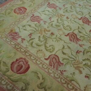 Floral vine rug in shades of cream, rose, and pale green, showing a decorative border on wood planks.