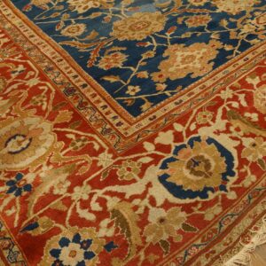 Blue and rust floral rug corner, showing detailed border patterns and beige fringe on wood floor.