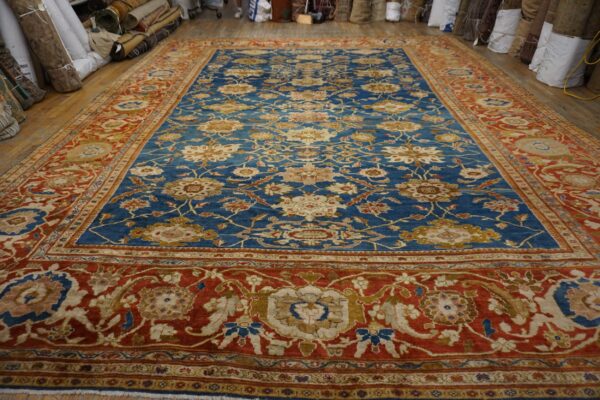 Large blue and red patterned rug with a floral motif displayed on a wooden floor near rolled rugs.