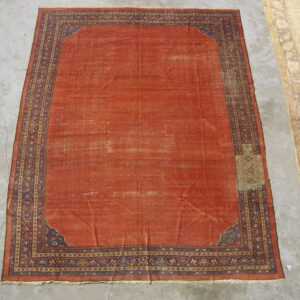 Distressed reddish-orange rug with a complex blue and gold geometric border on cracked concrete flooring.