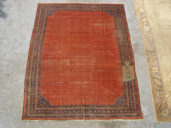 Distressed reddish-orange rug with a complex blue and gold geometric border on cracked concrete flooring.