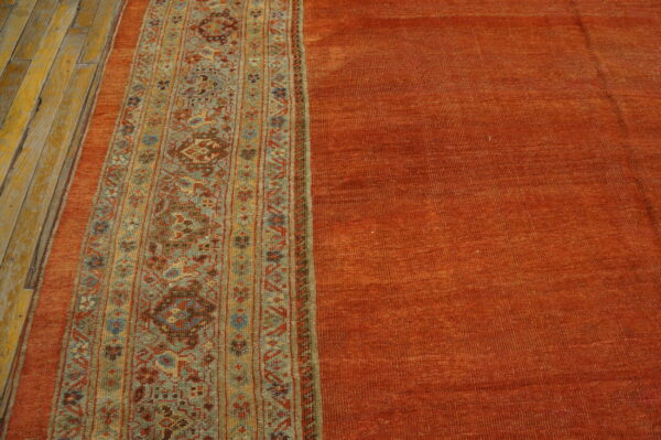 Textured orange rug with a patterned teal, gold, and red stripe placed next to worn yellow wood planks.
