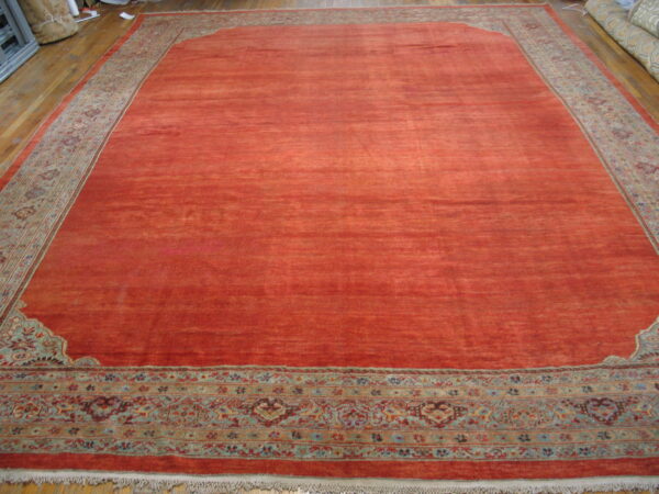 Large reddish-orange rug featuring a complex multicolored traditional border displayed on hardwood flooring.