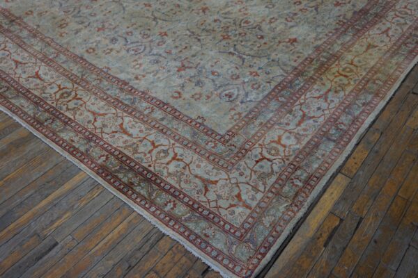 Ornate, low-pile rug in pale blue, beige, and rust colors rests on worn hardwood planks.