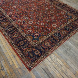 Intricate low-pile rug with a red field and navy border rests on worn plank wood flooring.