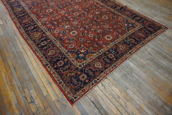 Intricate low-pile rug with a red field and navy border rests on worn plank wood flooring.