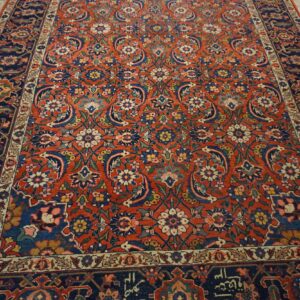 Ornate floral rug with a rust-red field and navy blue border on rustic wooden floor.