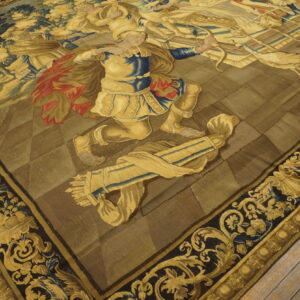 Detailed figural rug with gold, blue, and red figures against an olive background and ornate dark borders on wood flooring.