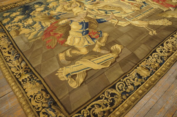 Detailed figural rug with gold, blue, and red figures against an olive background and ornate dark borders on wood flooring.
