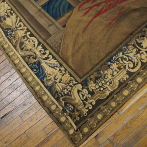 Muted brown and gold rug with an ornate scroll border on distressed wooden plank flooring.