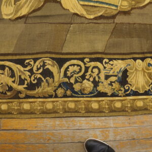 Khaki and brown woven textile with ornate dark gold borders resting on distressed wooden plank flooring.