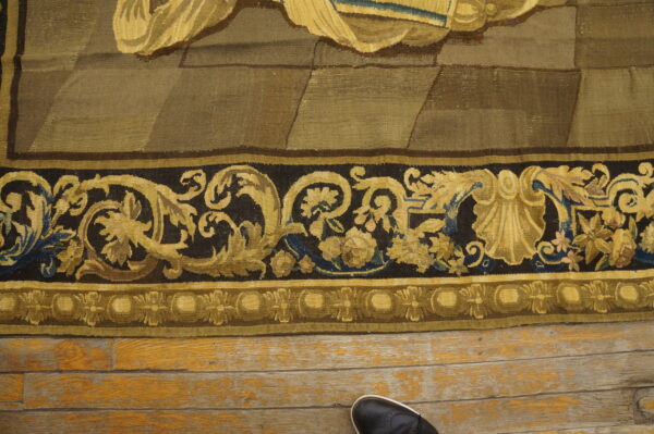 Khaki and brown woven textile with ornate dark gold borders resting on distressed wooden plank flooring.