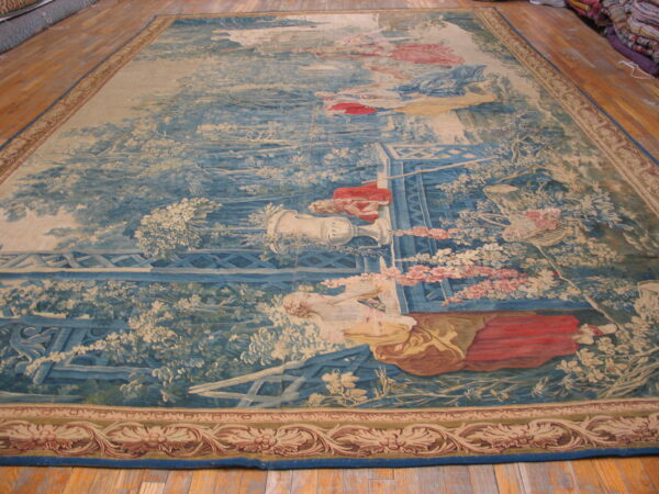 Detailed blue and beige tapestry rug depicts a pastoral garden scene with figures on wood plank floor.