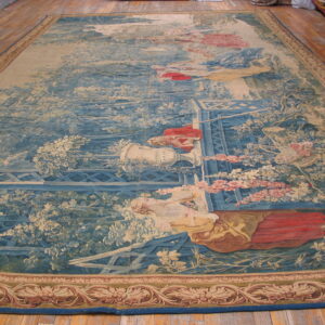 Pictorial rug showing figures and foliage in blue and beige tones, displayed on a wooden floor.