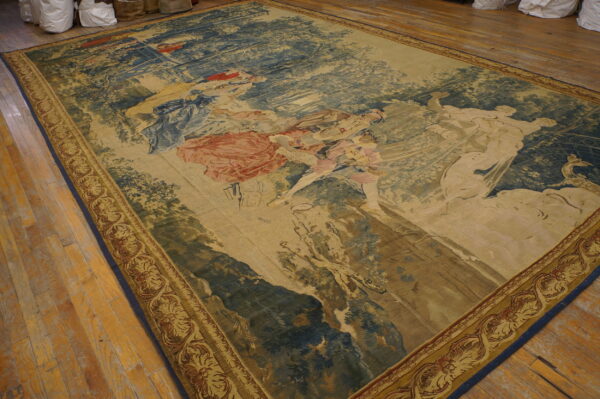 Large tapestry rug depicting classical figures in muted blue, tan, and red on worn wood flooring.