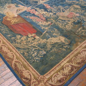 Low-pile tapestry rug featuring a figural garden scene in blue, green, red, and cream on distressed wood floors.