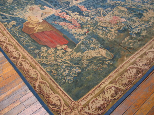 Low-pile tapestry rug featuring a figural garden scene in blue, green, red, and cream on distressed wood floors.