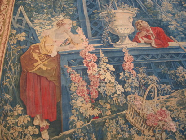 Historical tapestry with two figures in red and yellow leaning on a blue trellis, surrounded by pale pink flowers.