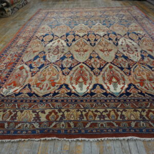 Ornate low-pile rug with red, navy, and cream floral lattice design rests on distressed wood plank flooring.