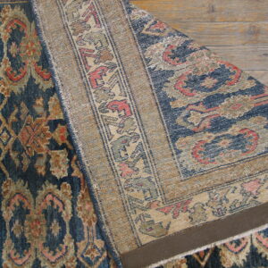 Low-pile blue and tan ornate rug partially folded to show the contrasting woven backing on wood floors.