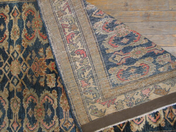 Low-pile blue and tan ornate rug partially folded to show the contrasting woven backing on wood floors.