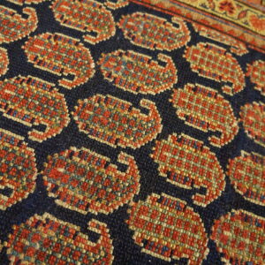 Close-up of low-pile rug with geometric red and gold motifs on navy blue, bordering a patterned stripe.