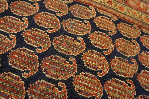 Close-up of low-pile rug with geometric red and gold motifs on navy blue, bordering a patterned stripe.