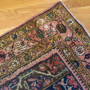 Corner view of a traditional rug featuring pink, navy, gold, and brown colors on wood floorboards.