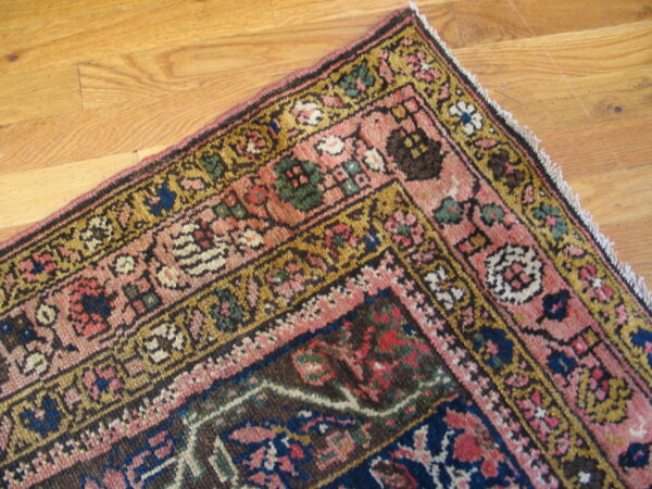 Corner view of a traditional rug featuring pink, navy, gold, and brown colors on wood floorboards.