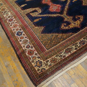 Corner view of a dark blue patterned rug with elaborate red and cream decorative borders on yellow-tinged wood.