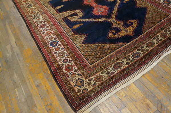 Corner view of a dark blue patterned rug with elaborate red and cream decorative borders on yellow-tinged wood.
