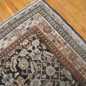 Ornate brown and beige patterned rug corner with complex borders on warm wooden plank flooring.