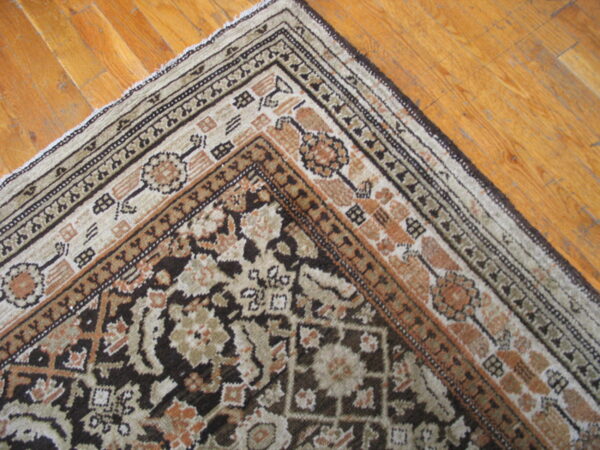 Ornate brown and beige patterned rug corner with complex borders on warm wooden plank flooring.