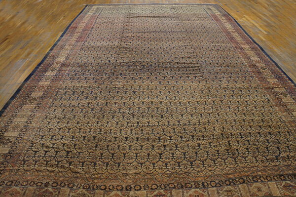 Oversize rug with tan and dark blue repeating geometric patterns displayed on light hardwood floor.