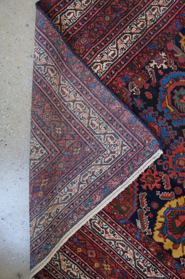 The woven underside of a navy, red, and beige patterned rug is visible as it is folded on concrete.