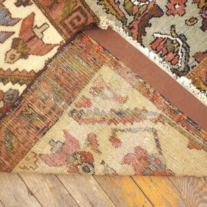 A multicolored rug is folded to reveal its woven backing, a brown pad, and distressed wood floorboards.