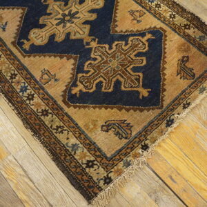 Diagonal view of a blue and tan geometric rug corner showing fringe on worn wooden floors.
