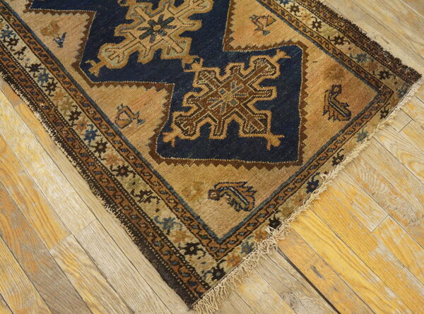 Diagonal view of a blue and tan geometric rug corner showing fringe on worn wooden floors.