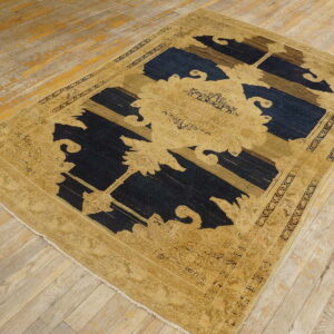 Tan and dark blue abstract rug with a stylized floral pattern rests on worn light hardwood flooring.