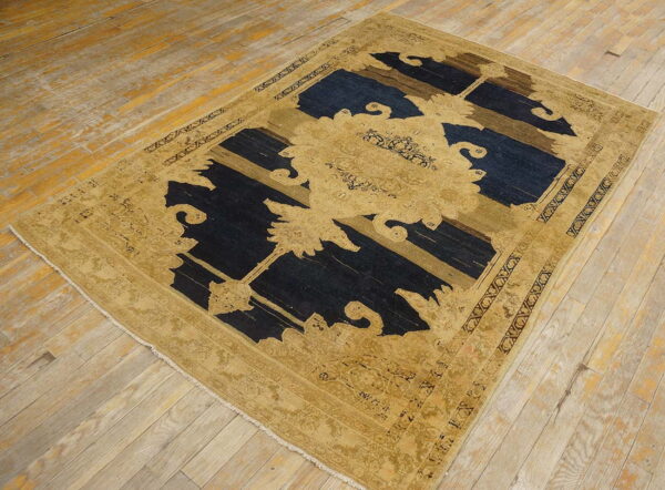 Tan and dark blue abstract rug with a stylized floral pattern rests on worn light hardwood flooring.