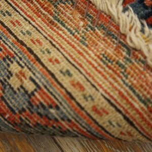 Close view of a rolled rug showing striped backing, cream fringe, and low pile blue and rust pattern.