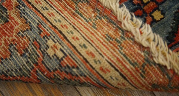 Close view of a rolled rug showing striped backing, cream fringe, and low pile blue and rust pattern.