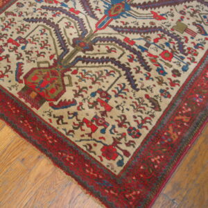A low-pile rug features a cream field, stylized red, blue, and green motifs, and a wide red border on wood plank flooring.