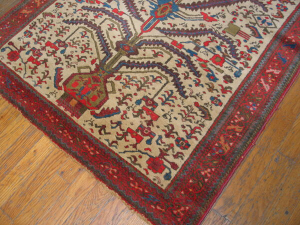 A low-pile rug features a cream field, stylized red, blue, and green motifs, and a wide red border on wood plank flooring.