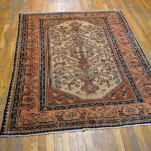 Low-pile rug featuring a central tree motif in tan, brown, and navy borders on worn hardwood flooring.