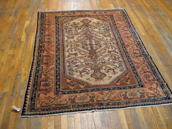 Low-pile rug featuring a central tree motif in tan, brown, and navy borders on worn hardwood flooring.