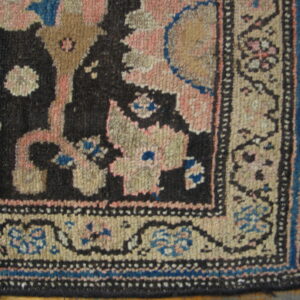 Close-up of a dark, rustic patterned rug corner featuring pink, tan, and blue on wood flooring.