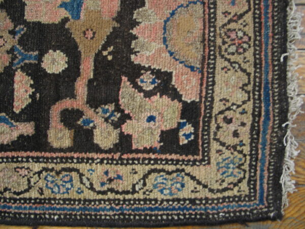 Close-up of a dark, rustic patterned rug corner featuring pink, tan, and blue on wood flooring.