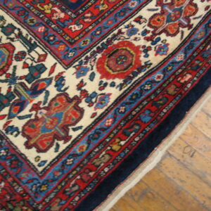 Traditional red, blue, and cream patterned rug corner placed over worn light brown wood flooring.